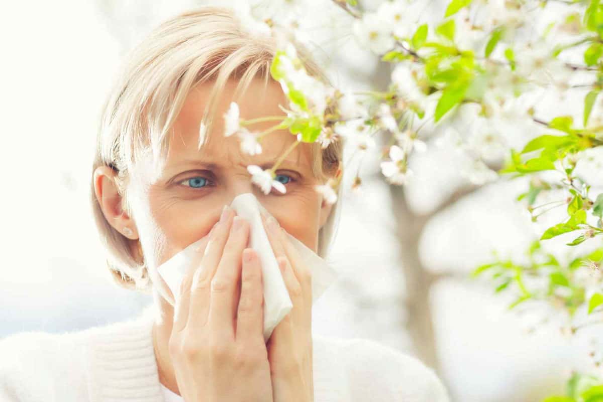 Bild von einer Frau vor einer Blüte mit Pollen, die sich mit einem Taschentuch schnäuzt, weil sie Heuschnupfen oder auch Rhinitis allergica bzw. allergischen Schnupfen hat